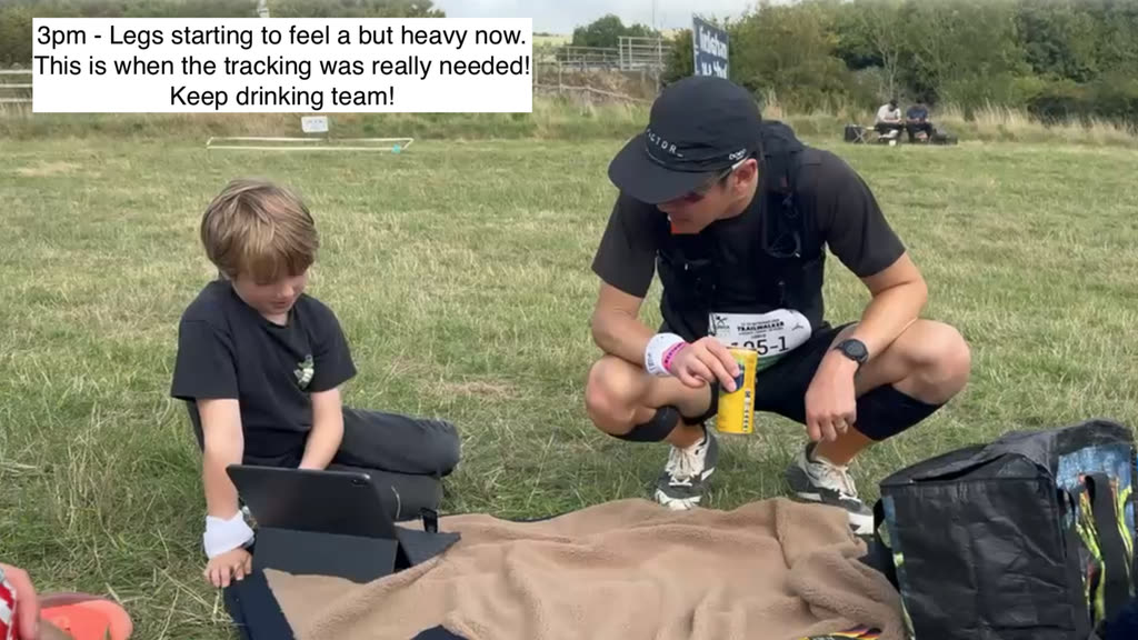 A boy sitting cross-legged on the grass with a tablet, beside a walker in a cap and race number crouching down with a drink.