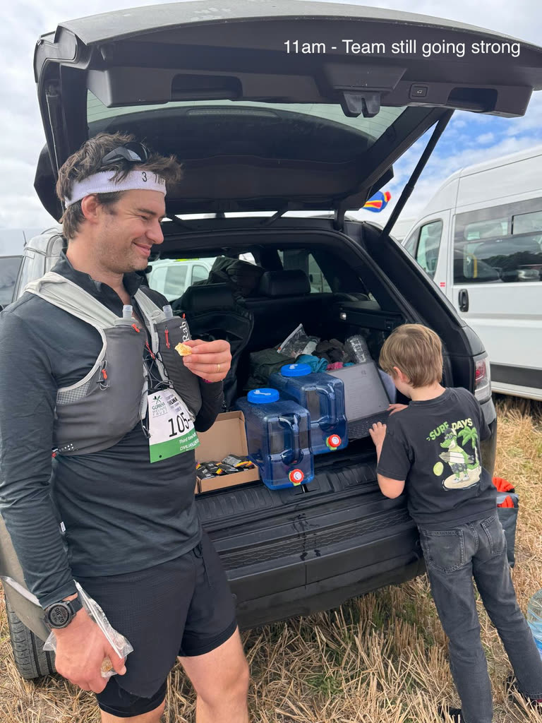 A walker in a race vest eating at the open boot of a car while a boy reaches into the boot past blue water carriers and a box of gels.