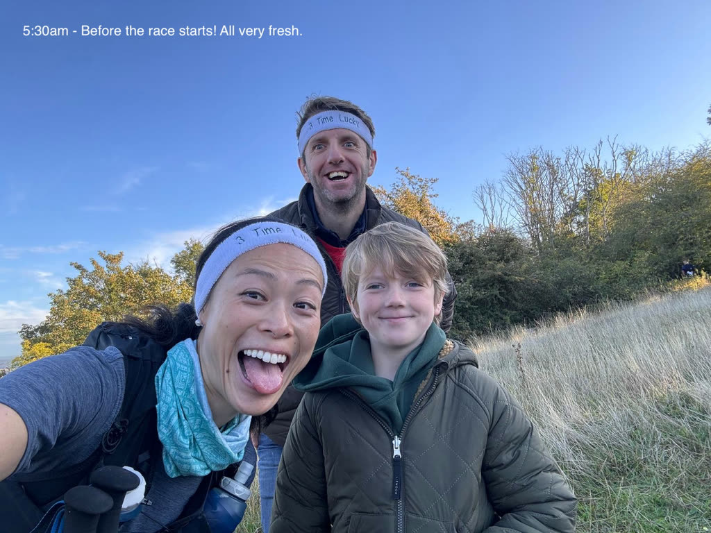 Three people smiling into the camera on a grassy hillside at dawn, two wearing white "3 Time Lucky" headbands, autumn trees and blue sky behind them.