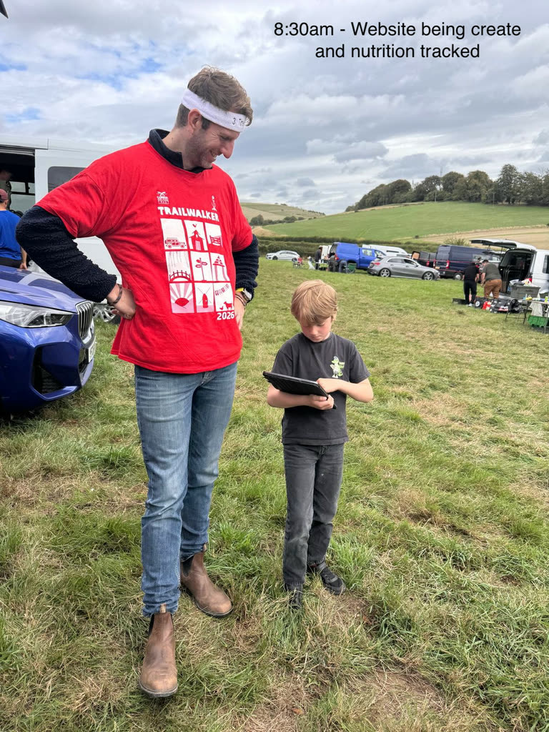 A boy standing on grass holding an iPad, concentrating on the screen, next to an adult in a red Trailwalker 2026 t-shirt and a white headband.